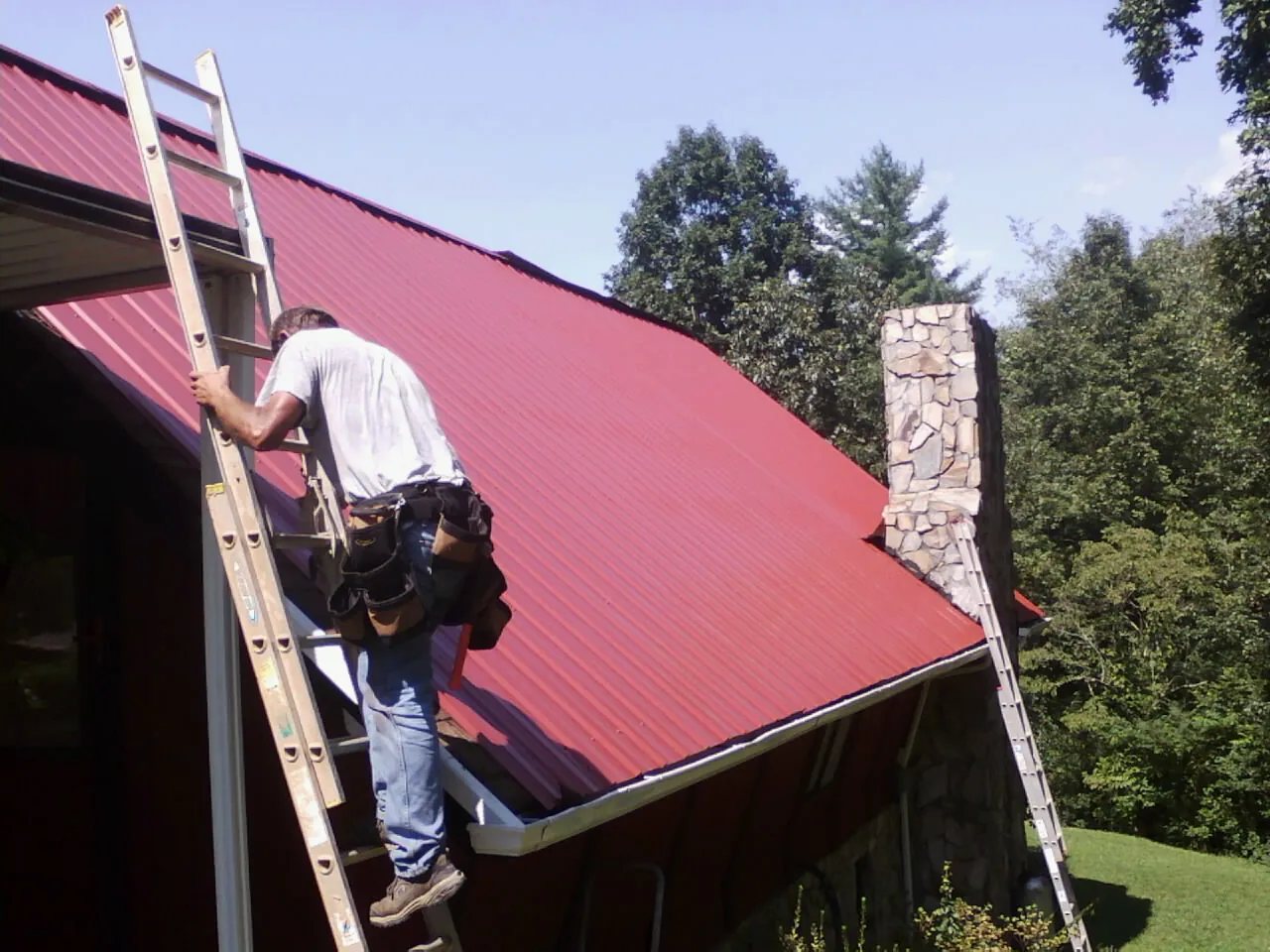 Roofer on a ladder beside a red metal roof and stone chimney — Soffit & Fascia Repair by Roofing Brothers in Wilder